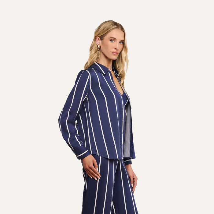 A woman with long blonde hair stands sideways, wearing the Ellery Shirt—a lightweight tencel button-down with vertical white stripes—paired with matching pants, against a plain light background.