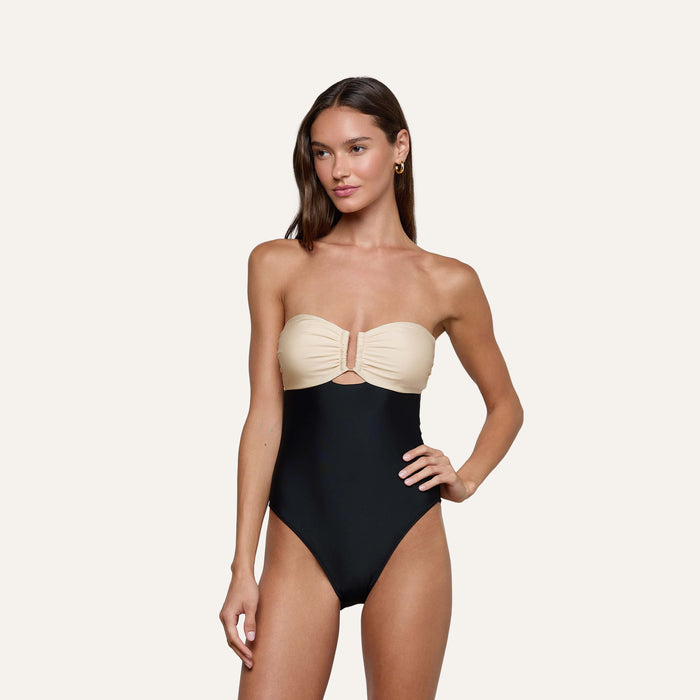 A woman with long brown hair poses against a plain light background, wearing the Emilia Bandeau One-Piece Swimsuit. The swimsuit features a beige bandeau-style top with ruched front detailing and a contrasting black high-leg bottom. She accessorizes her look with gold hoop earrings.