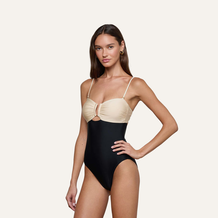 A woman stands against a plain light background wearing the Emilia Bandeau One-Piece Swimsuit, which features a color-blocked design with a beige top and black bottom, spaghetti straps, a high-leg cut, and a small cutout below the bust.