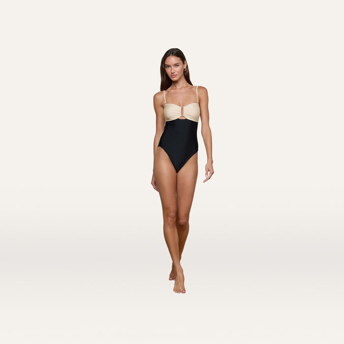 A woman stands barefoot against a plain white background, wearing the Emilia Bandeau One-Piece Swimsuit, which features a color-blocked design with a light beige top and black bottom. The high-leg cut accentuates her silhouette.