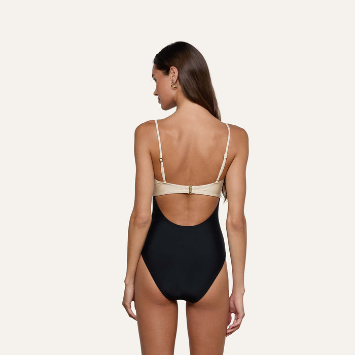 A woman with long brown hair wears the Emilia Bandeau One-Piece Swimsuit, a color-blocked black and beige bandeau style with a high-leg cut, standing with her back to the camera against a plain light background.