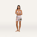 A woman stands barefoot with arms crossed, wearing a colorful bikini top and the Enyo Cover-Up Short, which features a high-waisted pull-on silhouette adorned with a vibrant postcard print of tropical sunsets and palm trees, against a plain white background.