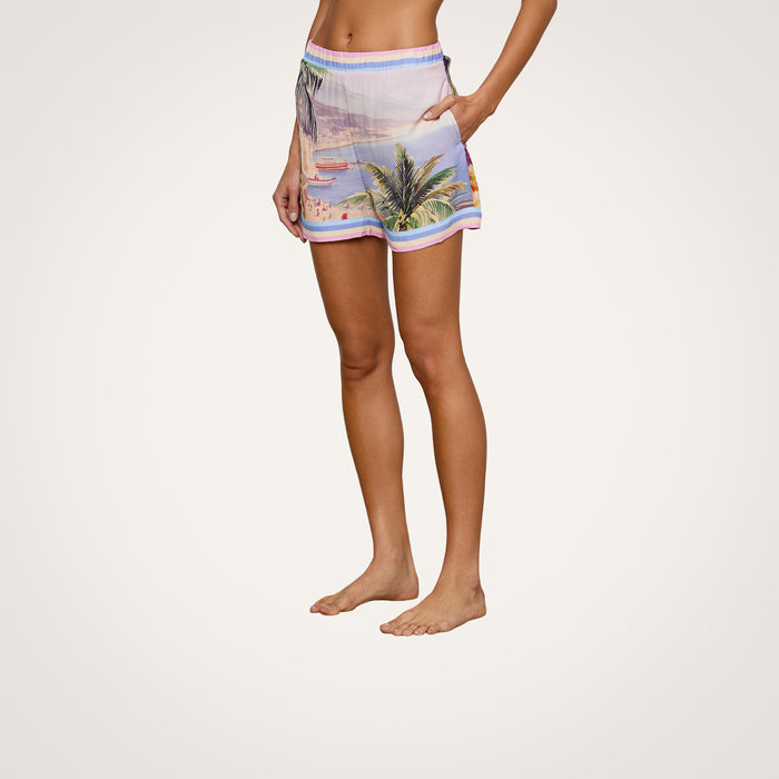 A person stands barefoot wearing the Enyo Cover-Up Short, which features a tropical beach print with palm trees and a sunset. The background is plain white, and only the lower torso and legs are visible.