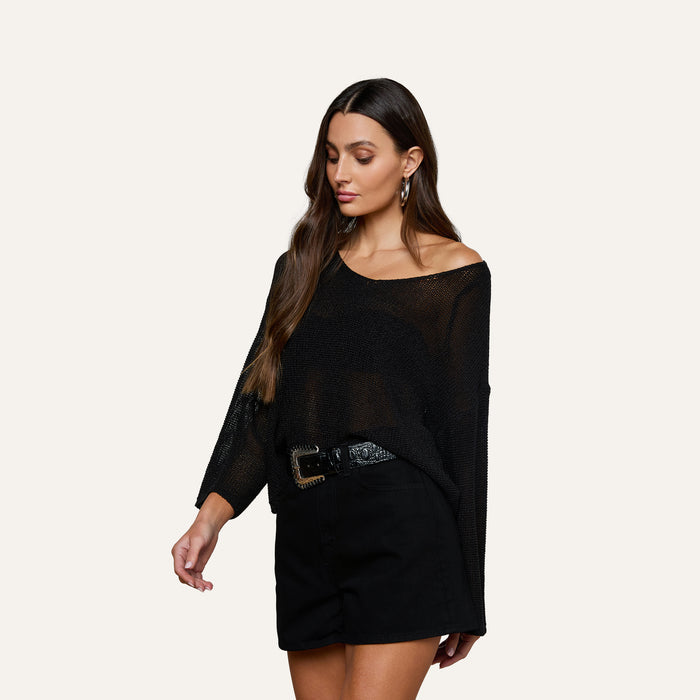 Woman wearing the Ethan Knit Pullover—a relaxed fit black knit top with an off-shoulder neckline—styled with black shorts, a black belt, and hoop earrings, posing against a plain light background.