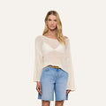 A woman wearing the Ethan Knit Pullover—a loose, off-white knit top with an off-shoulder neckline—paired with light blue denim shorts stands facing forward against a plain, light background. She has long blonde hair and a neutral expression.