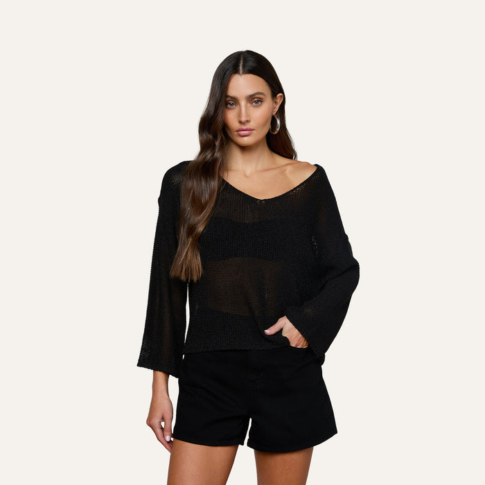 A woman with long wavy brown hair wears the Ethan Knit Pullover, a relaxed fit sheer black long-sleeve top, paired with black shorts. She has one hand in her pocket and stands against a plain light background, looking at the camera.