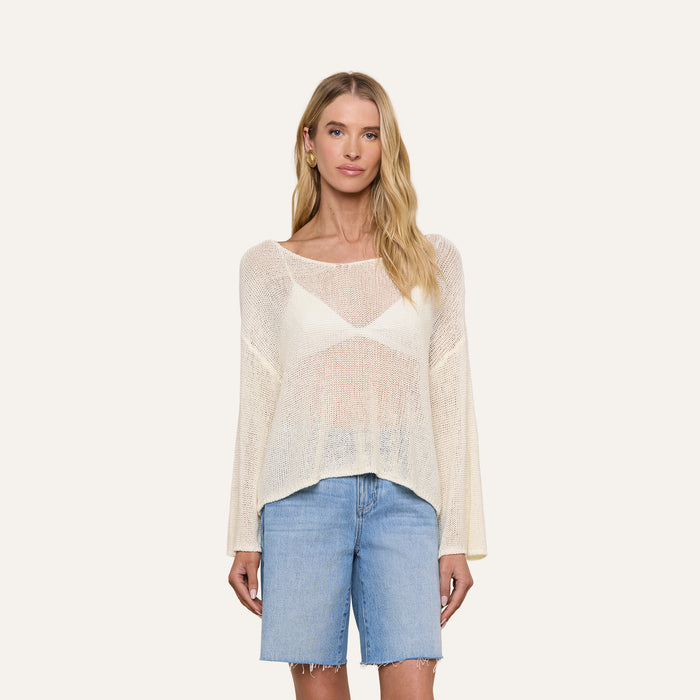 A woman stands facing forward against a plain light background, wearing the Ethan Knit Pullover—a sheer, cream, long-sleeve top with a relaxed fit—layered over a white bralette and paired with knee-length denim shorts.