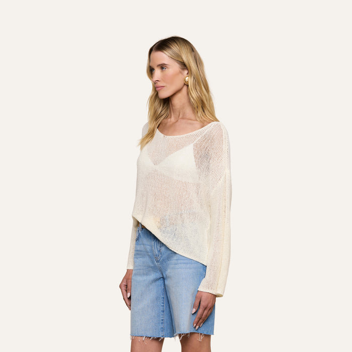 A woman with long blonde hair wears the Ethan Knit Pullover in a relaxed fit with an off-shoulder neckline over a white top and blue knee-length denim shorts, standing against a plain light background.