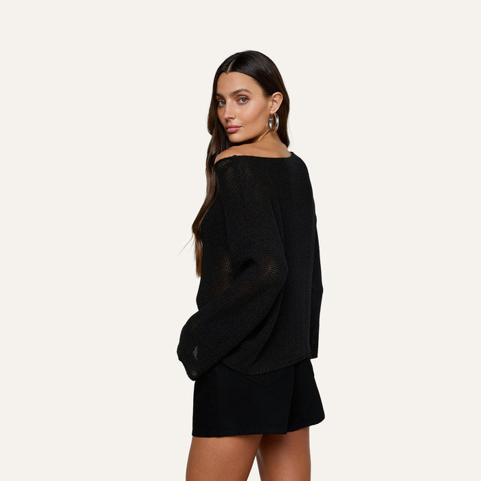 A woman with long brown hair, wearing hoop earrings and a relaxed fit Ethan Knit Pullover in black paired with black shorts, stands with her hands in her pockets, looking over her shoulder against a plain light background.