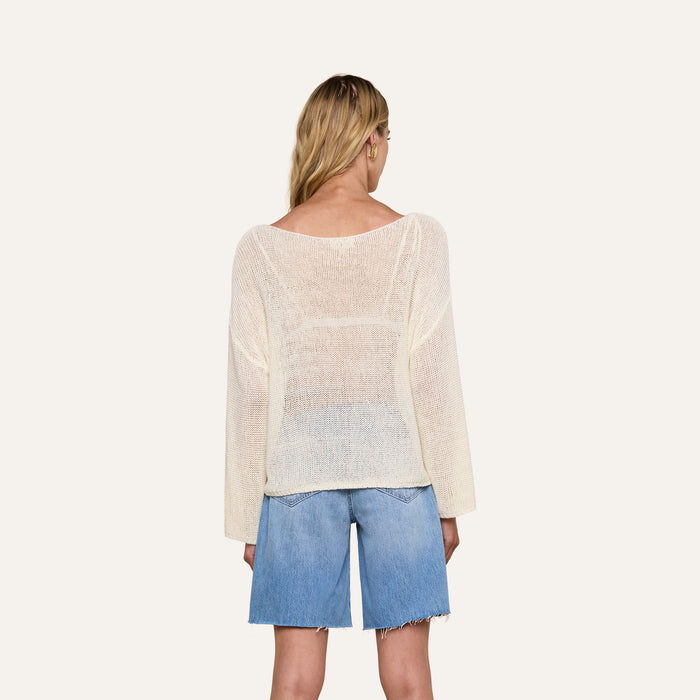 A woman with long blonde hair, seen from behind, wears the Ethan Knit Pullover with a relaxed fit and an off-shoulder neckline, paired with knee-length blue denim shorts against a plain light background.