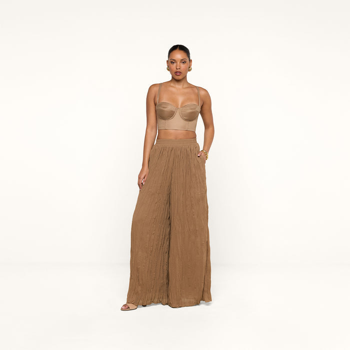 A woman stands against a plain white background wearing a beige crop top and the Ethel Pleated Pant, loose, high-waisted crinkle chiffon pants in brown, paired with sandals.