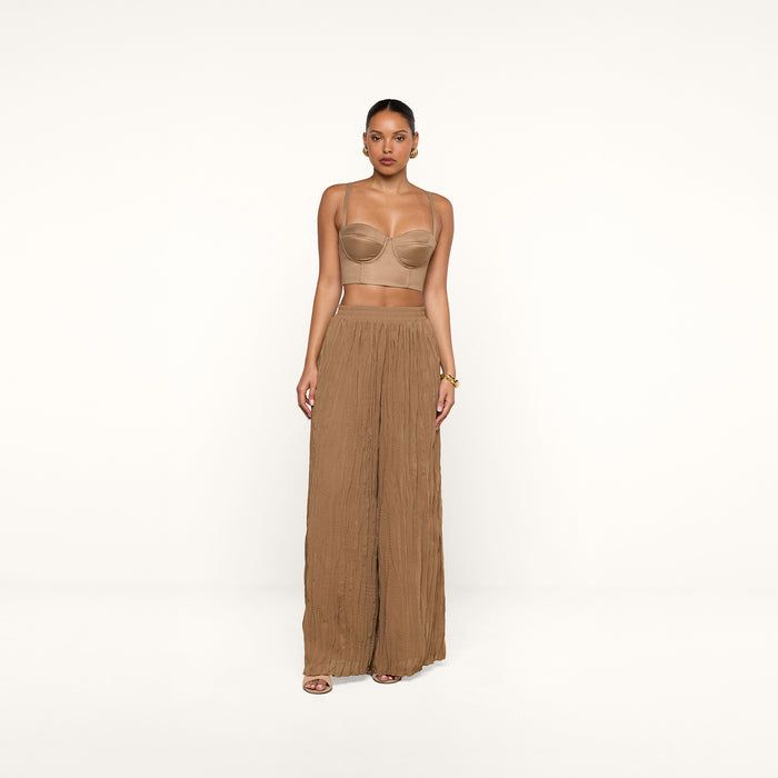 A woman stands against a plain white background wearing a tan bandeau top, the Ethel Pleated Pant—a sheer, wide-legged brown summer style—sandals, and gold hoop earrings. Her hair is pulled back and her hands rest relaxed at her sides.
