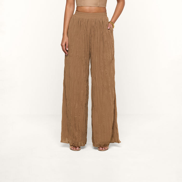 A person wearing the Ethel Pleated Pant, which features a high-waisted, wide-leg silhouette in light brown crinkle chiffon, and sandals stands against a plain white background. Their upper body is partially visible.