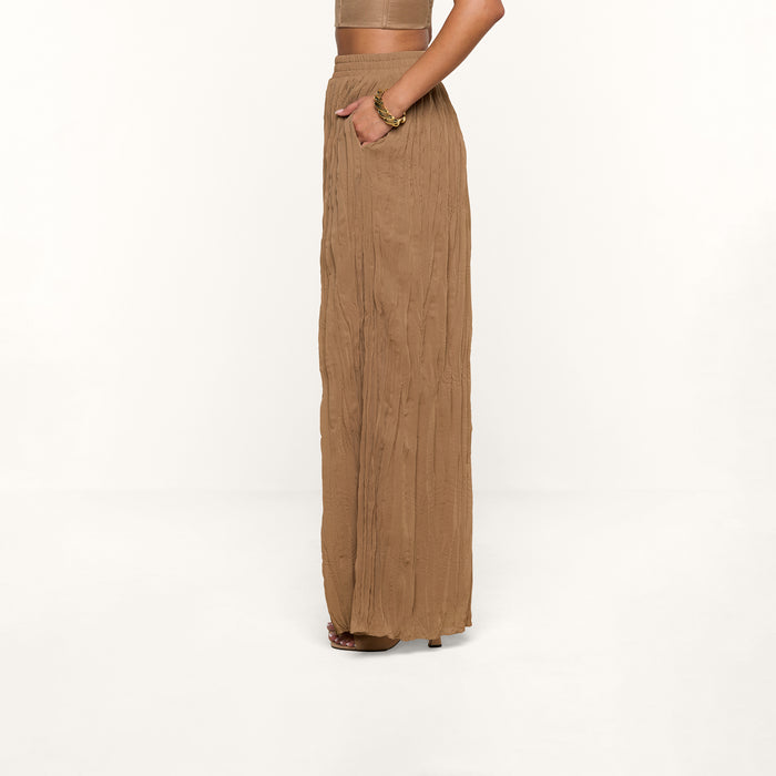A person stands sideways with hands in pockets against a white background, wearing a tan crop top and the high-waisted, loose, sheer Ethel Pleated Pant.