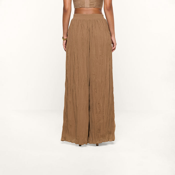 A woman stands facing away, wearing the Ethel Pleated Pant—a loose, high-waisted style in light brown crinkle chiffon—and a sleeveless cropped top against a plain white background.