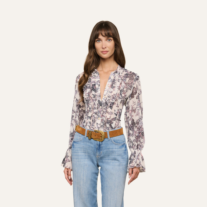 A woman wearing the Eunice Blouse, featuring a butterfly print and ruffled cuffs, paired with blue jeans and a tan belt, stands against a plain light background.