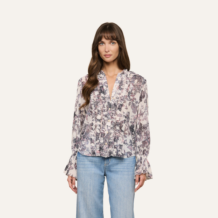 A woman is wearing the Eunice Blouse, a long-sleeve butterfly print top with ruffled cuff details, paired with light blue jeans and standing against a plain, light background.