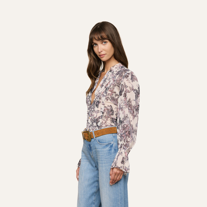 Woman with long brown hair stands against a plain background, wearing the Eunice Blouse—a butterfly print top—paired with blue high-waisted jeans and a tan belt. She looks slightly to her left with a relaxed expression, her hands by her sides.