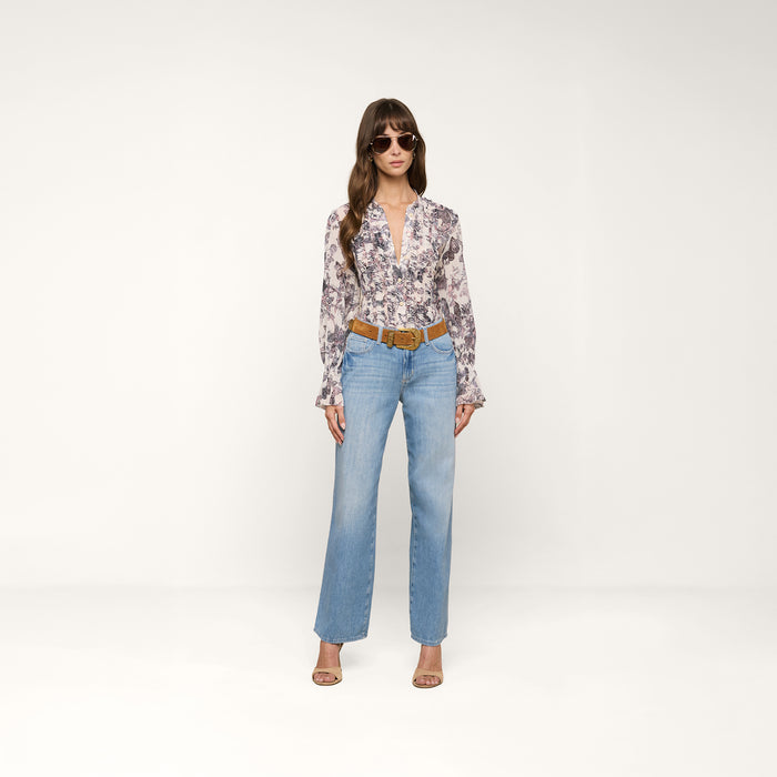 A woman wearing sunglasses, the Eunice Blouse with a butterfly print and long sleeves, light blue jeans, a brown belt, and sandals stands against a plain white background.