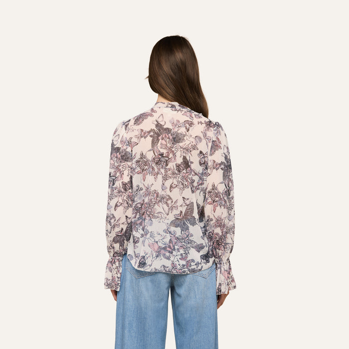 A woman with long brown hair is shown from the back against a plain light background, wearing the Eunice Blouse featuring a butterfly print and paired with light blue jeans.