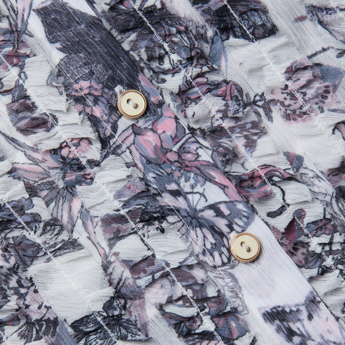 Close-up of the Eunice Blouse fabric featuring a pink, purple, and gray floral pattern, with two beige buttons bordered by gold rims sewn onto it. The textured, slightly sheer material showcases visible stitching lines—details reminiscent of those found on ruffled cuffs or butterfly print blouses.
