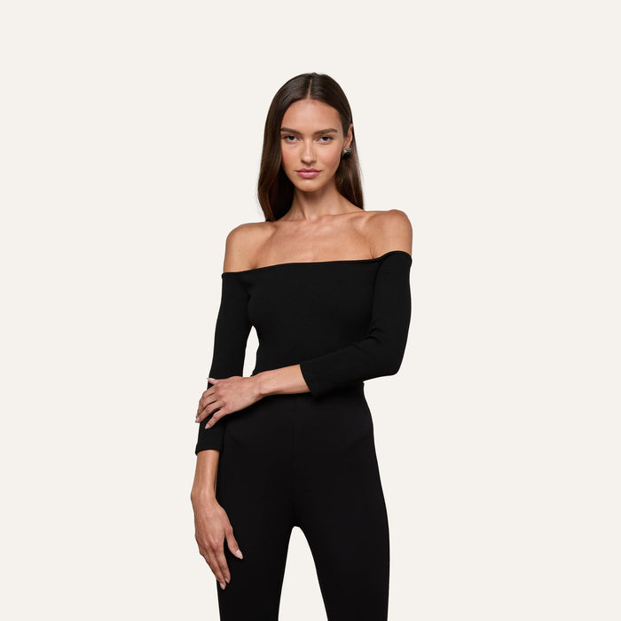 A woman stands against a plain light background, dressed in the Evelyn Off-The-Shoulder Bodysuit—an off-the-shoulder black piece with three-quarter sleeves—paired with matching black pants. Her long straight brown hair frames her neutral expression, and the bodysuit is made from stretch cotton modal for a comfortable fit.