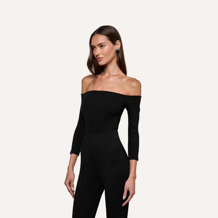 A woman stands against a plain light background, looking to the side, wearing the Evelyn Off-The-Shoulder Bodysuit in stretch cotton modal with three-quarter sleeves and black pants.