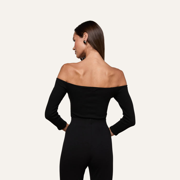 The woman, facing away from the camera, wears the Evelyn Off-The-Shoulder Bodysuit—crafted from stretch cotton modal—in black, paired with black pants as she stands against a plain light background.