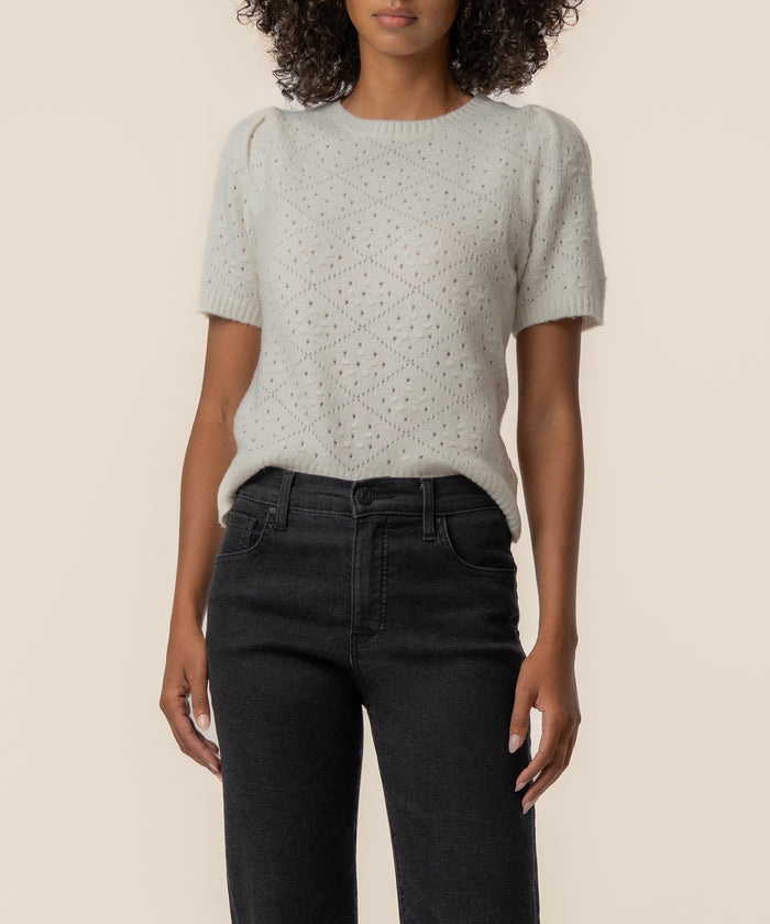 Cream knit sweater with textured diamond pattern and short sleeves worn with black jeans.