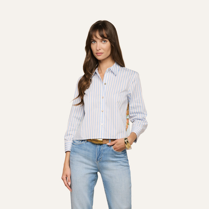 A woman wearing the Fawn Beach Print Shirt—an organic cotton button-up with light blue and white stripes—and light blue jeans stands against a plain background, looking at the camera with one hand in her pocket.
