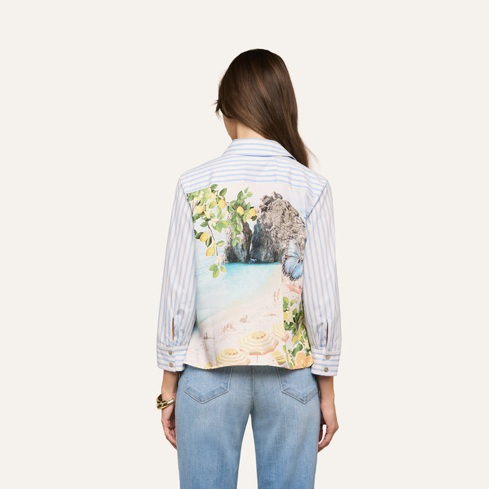 A woman with long brown hair, wearing blue jeans and the Fawn Beach Print Shirt—an organic cotton shirt featuring a colorful beach scene on the back—stands facing away from the camera.