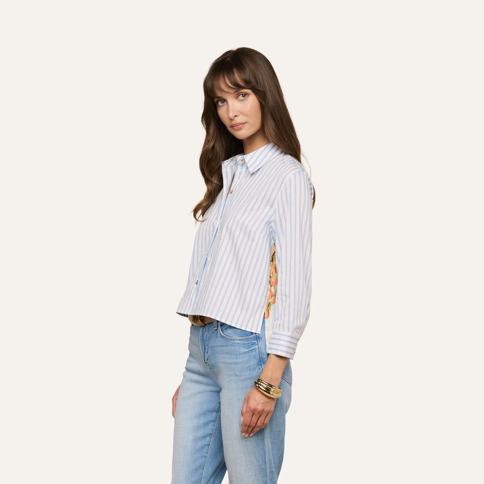 A woman with long brown hair is wearing the Fawn Beach Print Shirt—an organic cotton, blue and white striped shirt featuring a floral back detail—paired with blue jeans and gold bracelets as she stands against a plain light background.