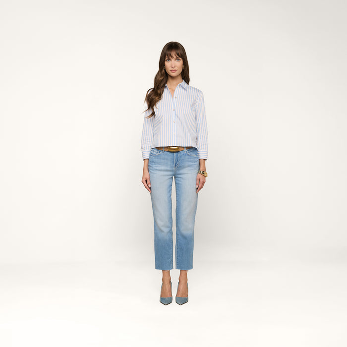 A woman with long brown hair stands facing forward in a white studio, wearing the Fawn Beach Print Shirt—crafted from organic cotton and featuring blue-and-white stripes—paired with light blue jeans, blue heels, and a brown belt. Her hands rest by her sides.