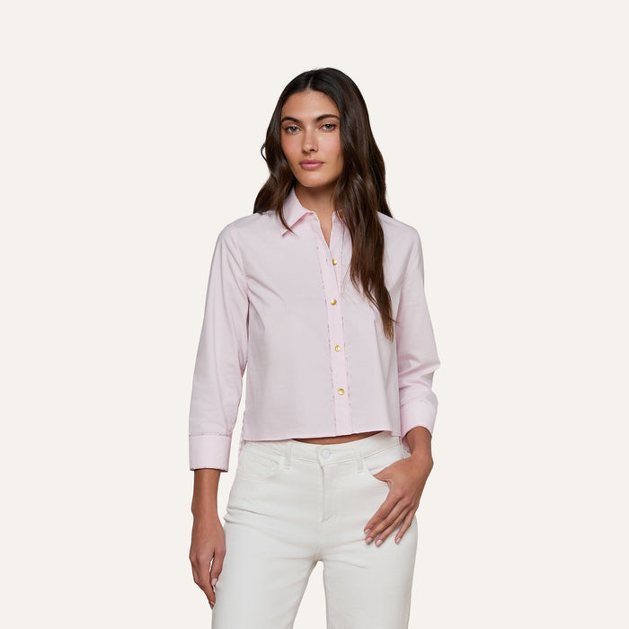 A woman models the Fawn Shirt, a cropped pale pink top, paired with white pants as she stands against a plain, light background with one hand in her pocket.