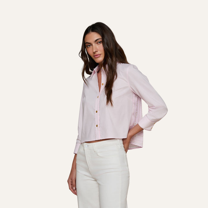 A woman with long brown hair wears the Fawn Shirt—a cropped pale pink top—paired with high-waisted white pants, standing against an off-white background and looking at the camera.