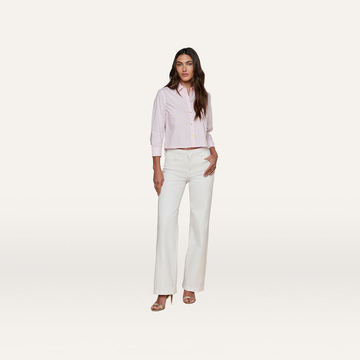 A woman stands against a plain white background, wearing the cropped pale pink Fawn Shirt paired with white high-waisted wide-leg pants and heeled sandals. She has long brown hair and one hand in her pocket.