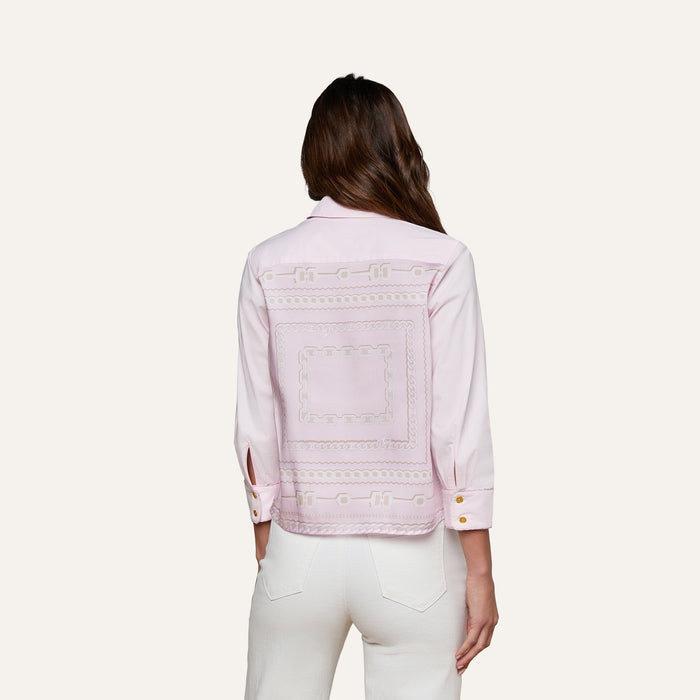 A woman with long brown hair wears the Fawn Shirt, a cropped pale pink top with an embroidered square design on the back, paired with white pants. She stands facing away against a plain, off-white background.