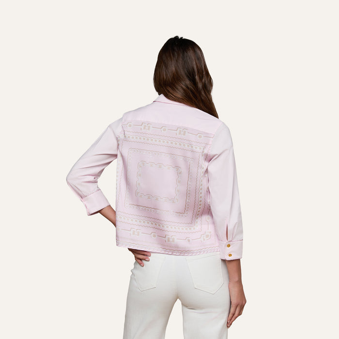 A woman with long brown hair, standing with one hand in her pocket and facing away from the camera against a plain background, wears the Fawn Shirt—a cropped pale pink top featuring embroidered patterns on the back—paired with white pants.