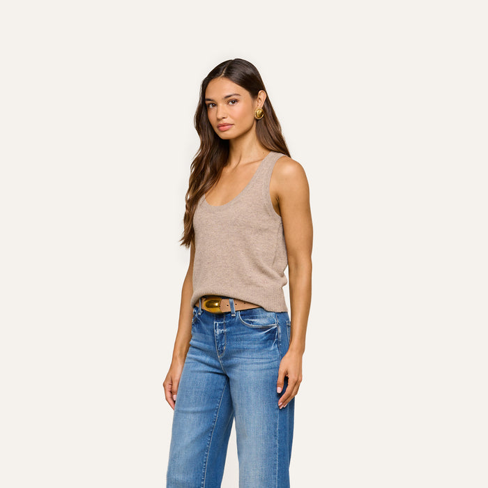 A woman with long brown hair wears the Filou Cashmere Tank—a sleeveless, lightweight beige top—paired with blue jeans, a brown belt, and gold hoop earrings, standing against a plain light background.