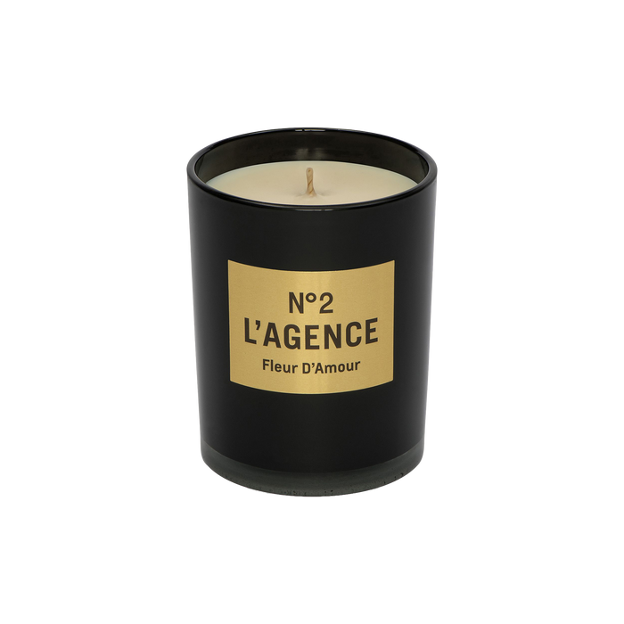 A black candle jar with a beige label displaying “Fleur D’Amour,” containing an unlit, cream-colored soy-blend wax candle infused with baccara rose.