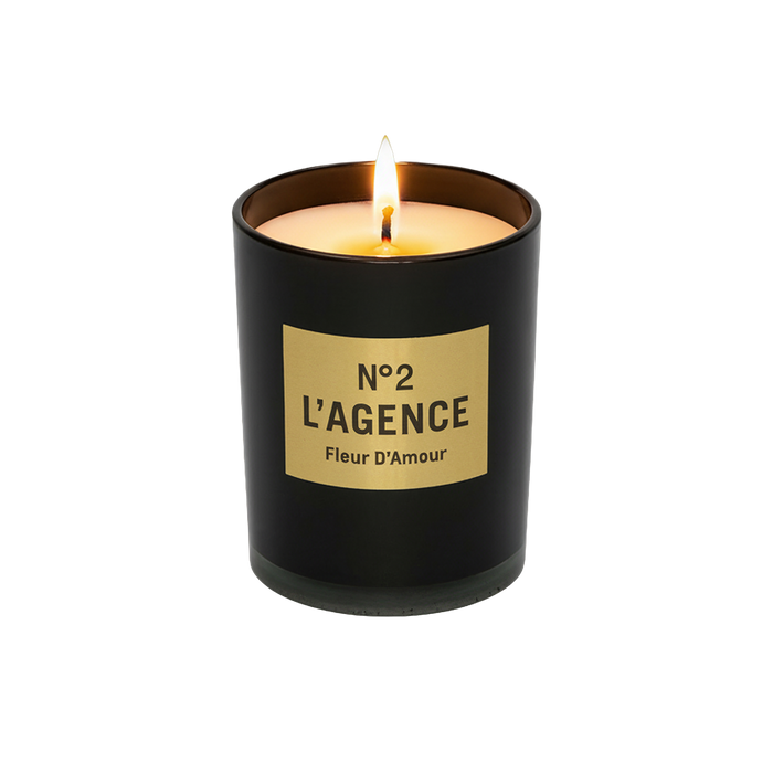 A lit Fleur D’Amour soy-blend wax candle in a black glass holder, featuring baccara rose notes, displayed on a plain white background.