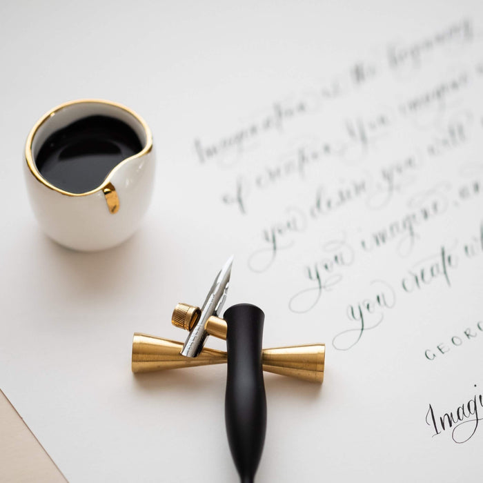 The Flourish calligraphy pen with oblique nib setting in Black on a brass pen rest on top of a sheet of modern calligraphy with an ink well