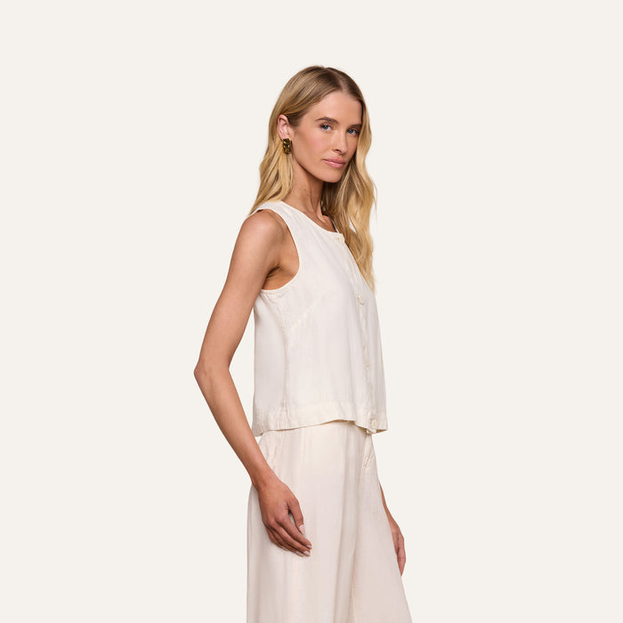 Wearing the Foster Linen Top, a woman with long blonde hair stands sideways against a plain white background, pairing the sleeveless linen top with a matching cream A-line skirt.