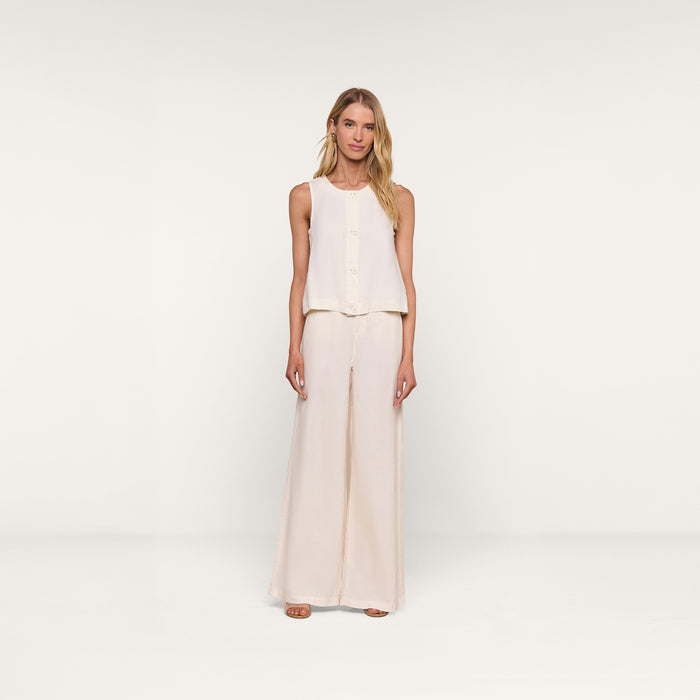 A woman stands against a plain white background, wearing the sleeveless Foster Linen Top with an A-line silhouette and matching wide-leg pants, paired with open-toe shoes.