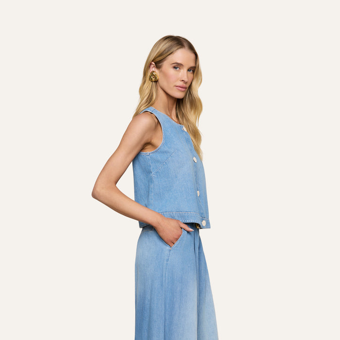 A woman with long blonde hair wears the sleeveless Foster Top in featherweight denim and matching wide-leg denim pants, standing in profile with her hands in her pockets against a plain light background.