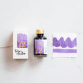 Tom's Studio Lavender Fountain Pen Ink – bottle with gold lid, boxed packaging featuring a purple and gold label marked '22', and a swatch of the vibrant lavender ink on textured paper.