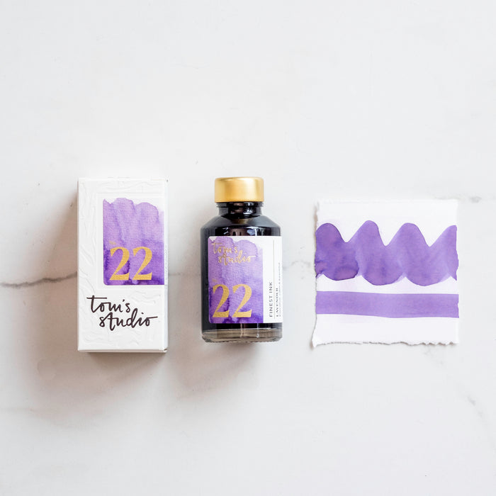Tom's Studio Lavender Fountain Pen Ink – bottle with gold lid, boxed packaging featuring a purple and gold label marked '22', and a swatch of the vibrant lavender ink on textured paper.
