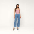 A woman stands against a plain white background wearing a pink tank top, the Frasier Rodeo Jean—a high-rise, wide-leg denim style with a slouchy fit—and blue heels. She has long brown hair and a neutral expression.