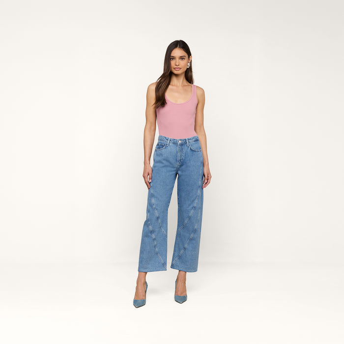 A woman stands against a plain white background wearing a pink tank top, the Frasier Rodeo Jean—a high-rise, wide-leg denim style with a slouchy fit—and blue heels. She has long brown hair and a neutral expression.