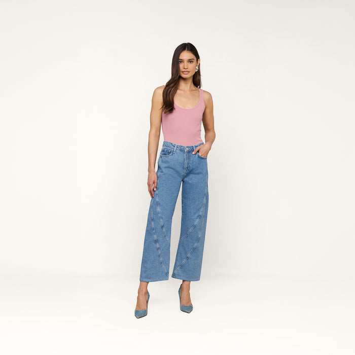 A woman with long, straight brown hair and a neutral expression stands against a plain background, wearing a pink sleeveless top, blue heels, and the high rise Frasier Rodeo Jean in a wide-leg blue denim style.